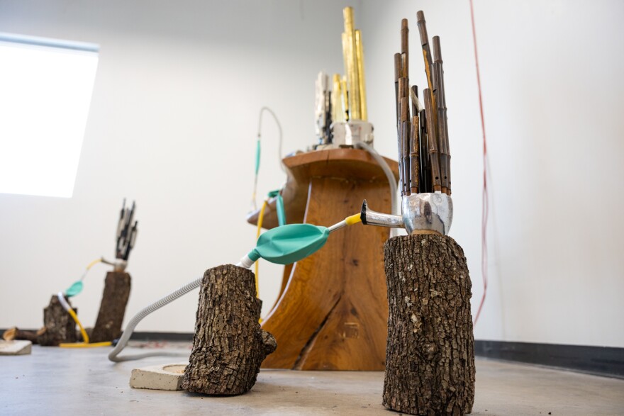 Small tree trunks sit on the floor of an art exhibit with a hand made instrument sitting on top consisting of a bag and tubes. 