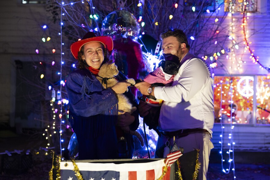 A woman with a red cowboy hat holds a cat next to a man holding a small dog as they hold the animals' paws out toward each other. In the background are holiday lights. 