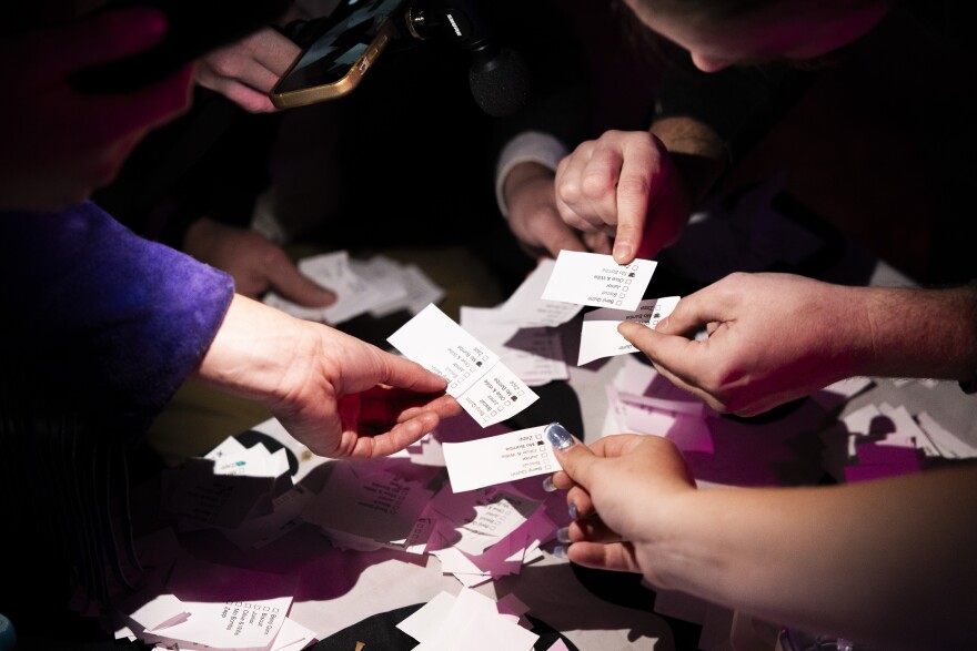 Hands are shown above a table holding pieces of paper with votes on them. On the table are more ballots. A person at top leans in to look closely at a vote. 