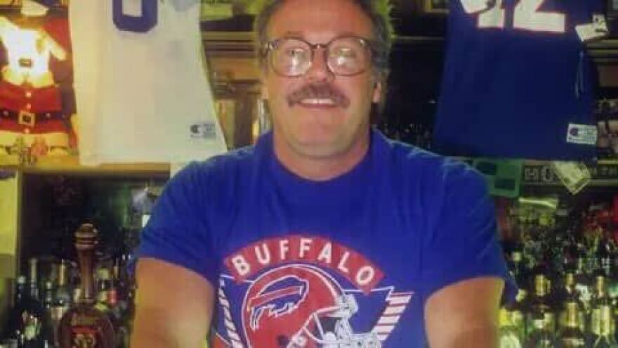 A man shown from the chest up smiling in front of bottles of liquor.