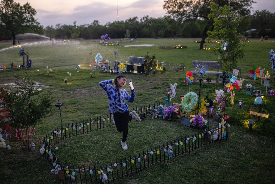 A young girl is seen dancing at a gravesite enclosed with a small fence and adorned with wreaths and flowers.