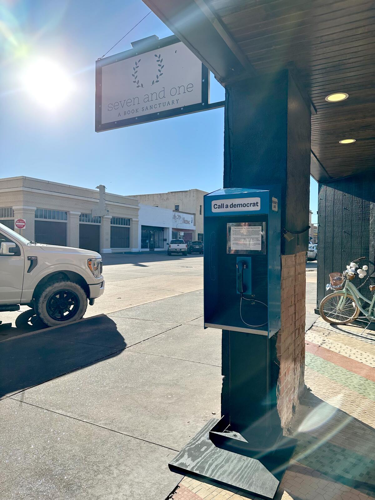 A pay phone rests against a pillar beneath a store sign on a street in a small city.