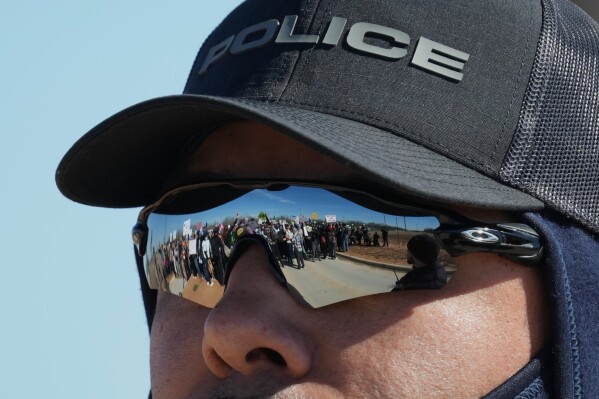 A police officer watches protesters, reflected in his sunglasses, gathered outside the South Texas Family Residential Center detention facility where Liam Ramos and his father are being detained in Dilley, Texas, Wednesday, Jan. 28, 2026. (AP Photo/Eric Gay)