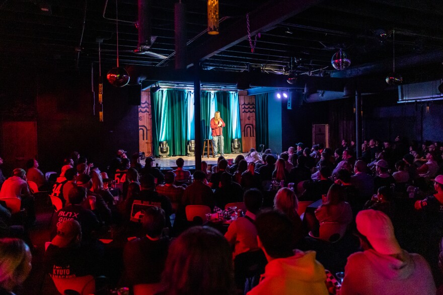 A man is shown standing on stage in the background with the audience shown in the foreground.
