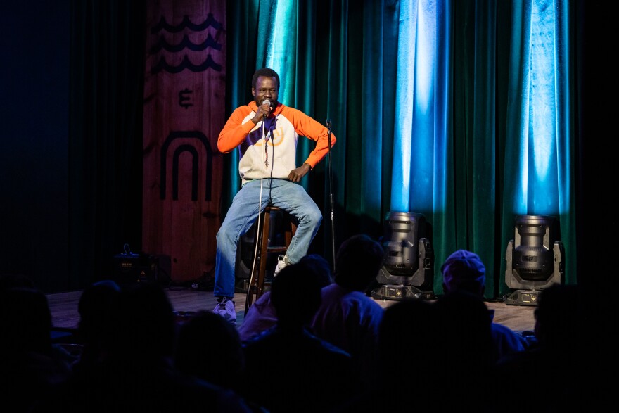 A man sits on a stool on stage holding a microphone in front of an audience who is shown at the bottom of the image.