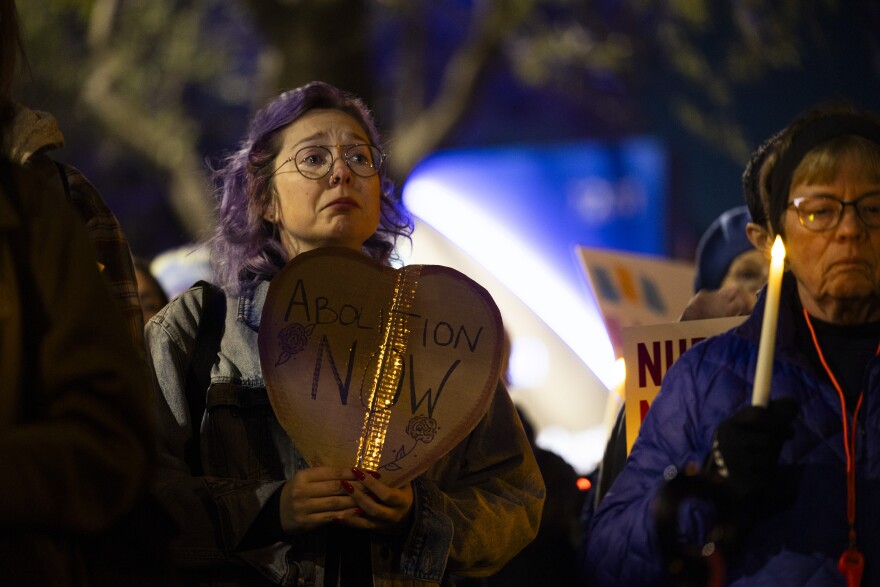 A woman with purple hair holds a heart shaped sign reading Abolition Now.