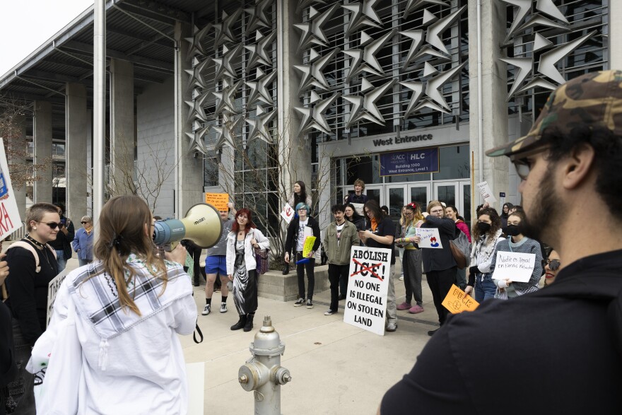 Protesters gather outside a building. One person has a sign that reads "Abolish ICE! No one is illegal on stolen land."