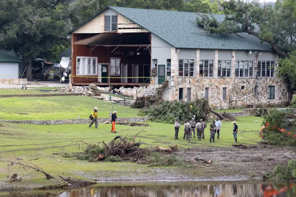 Law enforcement agents stand outside a damaged building which is missing a wall. In the foreground part of a river can be seen. 