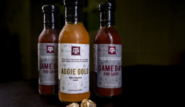 Bottles of Aggie Gold and Game Day BBQ sauce against black background with Texas A&M senior rings