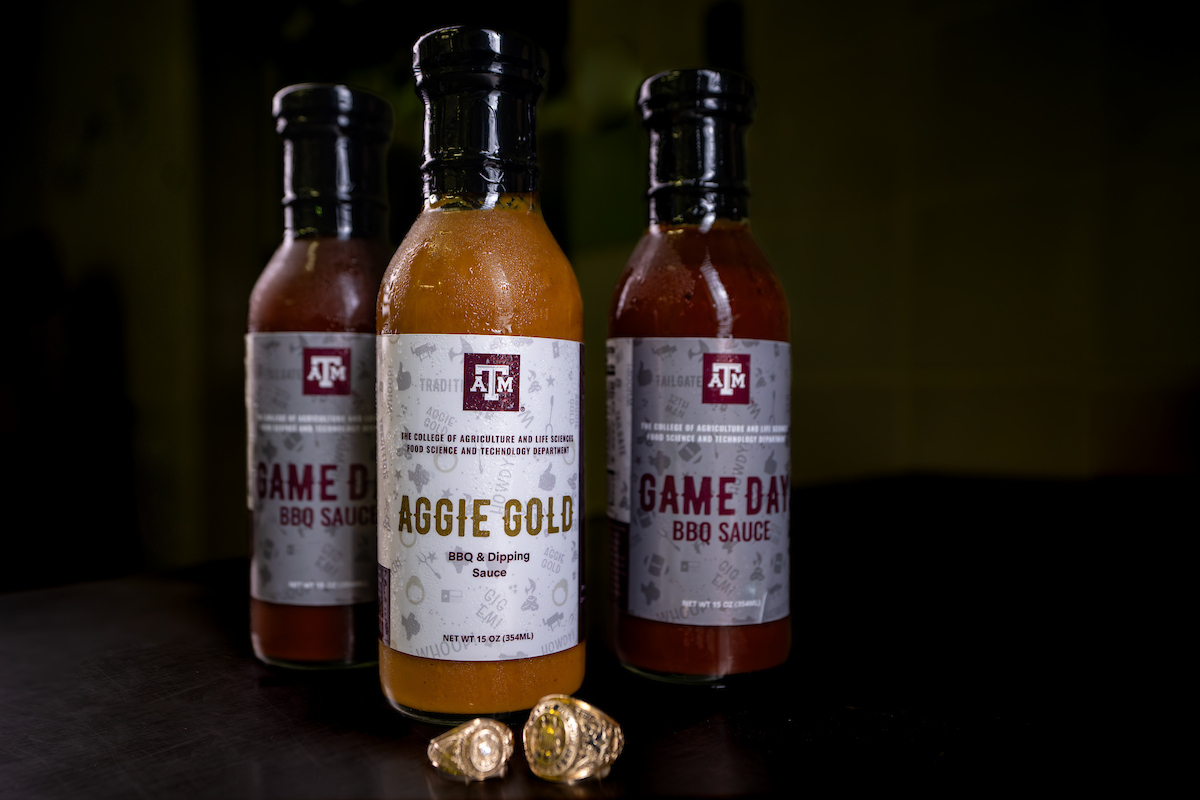 Bottles of Aggie Gold and Game Day BBQ sauce against black background with Texas A&M senior rings