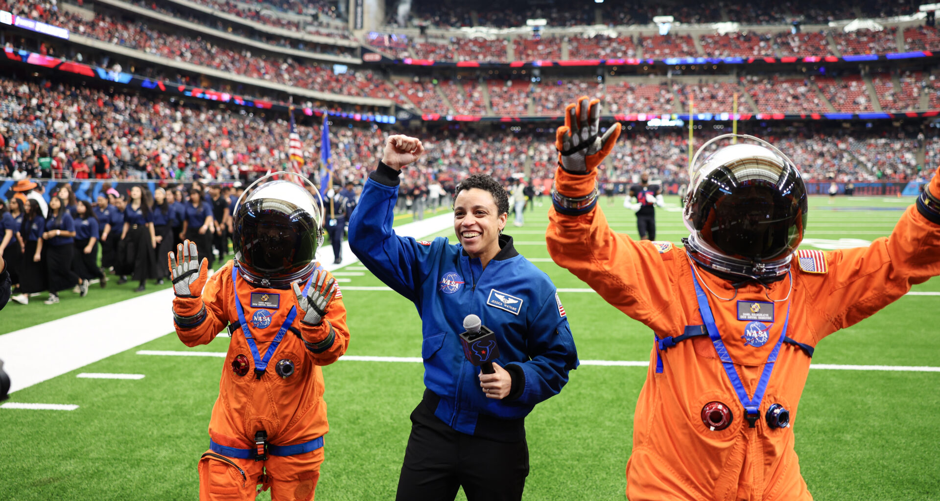 Three people pose with their hands in the air on the football field. Two people are wearing an orange spacesuit and the person in the middle is wearing a blue flight jacket.