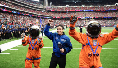 Three people pose with their hands in the air on the football field. Two people are wearing an orange spacesuit and the person in the middle is wearing a blue flight jacket.