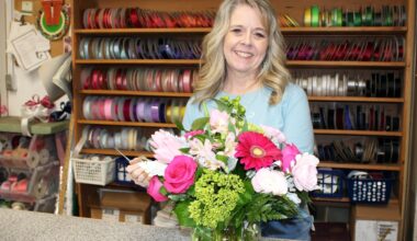 Female standing behind a flower arrangement on a counter in front of shelves holding spools of ribbon.