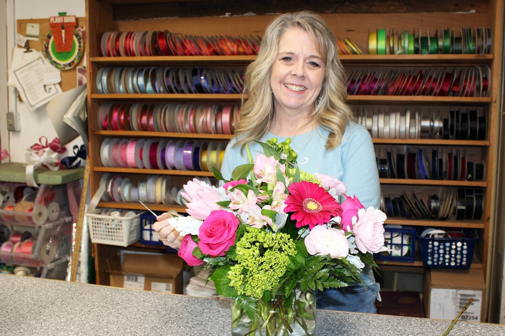 Female standing behind a flower arrangement on a counter in front of shelves holding spools of ribbon.