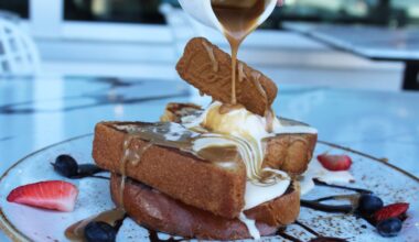 French toast on a plate topped with ice cream and a cookie. Liquid topping is being poured on the toast.