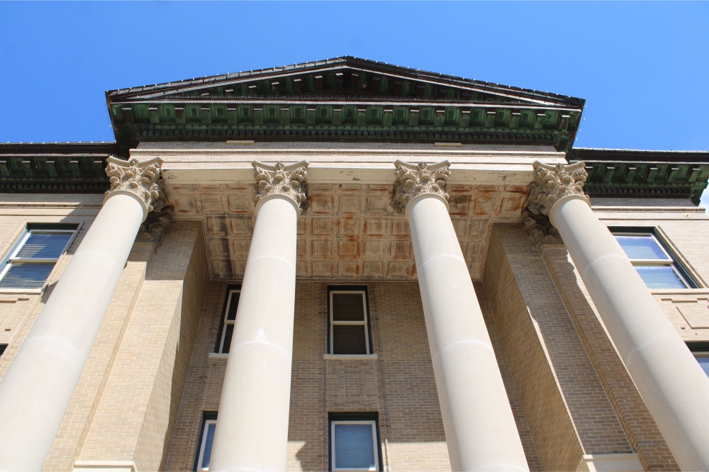 The Hays County courthouse, four large white pillars lead to the green, triangular roof.