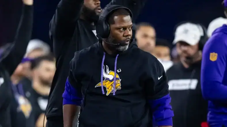 Minnesota Vikings coach, Daronte Jones, wearing a black hoodie and headset stands on the sideline during a game, looking toward the field with players and staff in the background.