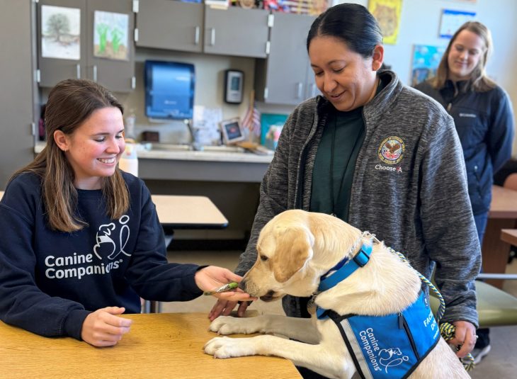 A smiling woman in a "Canine Companions" shirt holds a treat for a yellow lab in a service vest. Another woman and a person in the background watch.