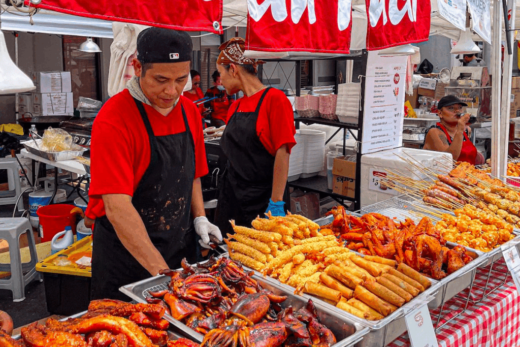 Image shows a vendor at an Asian food festival in front of trays of Asian food.