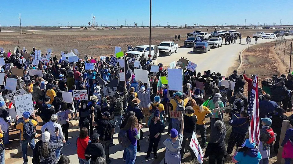 dozens of protesters hold up signs on a desert road as officers' vehicles sit in the distance