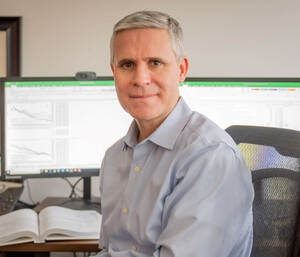 Middle-aged man with gray hair and a light blue shirt smiles at the camera. He sits in an office with two monitors displaying charts and an open textbook on his desk.