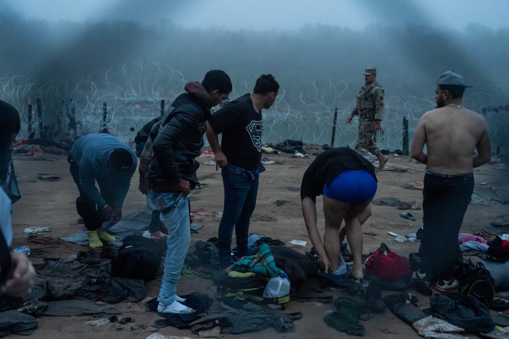 Migrants change clothes on the river bank, while a U.S. authority figure stands nearby, with razor wire in the background.
