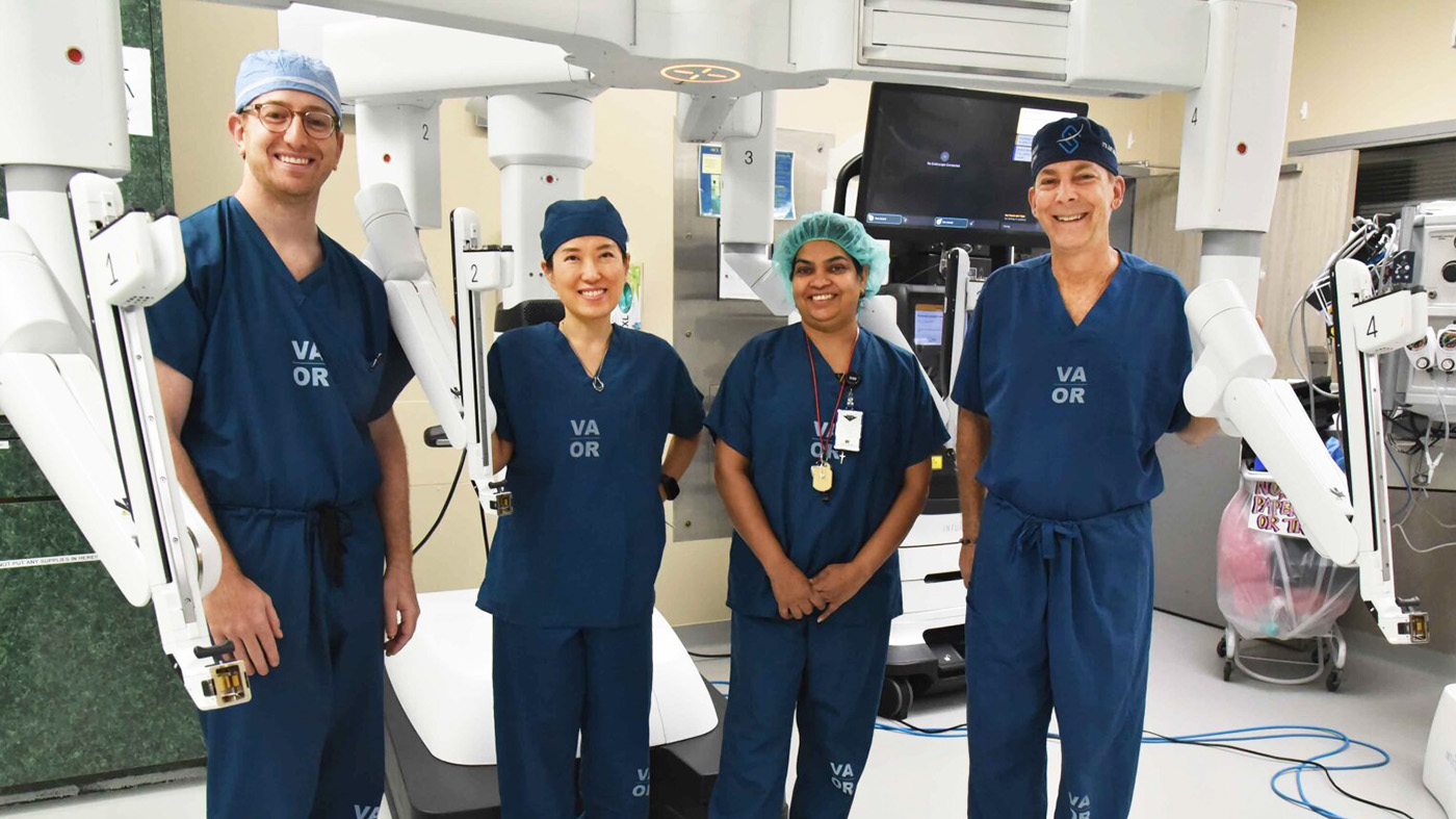 A diverse surgical team stands proudly in an operating room, wearing blue scrubs. They are posed in front of advanced robotic surgical equipment, smiling confidently.