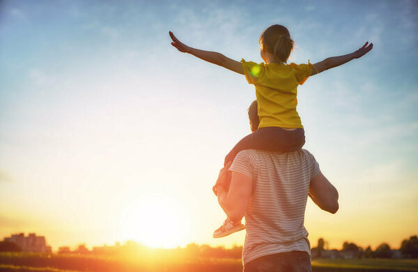 A young girl in a yellow shirt with a ponytail rides on an adult's shoulders, arms outstretched like wings, facing a vibrant orange and blue sunset sky.