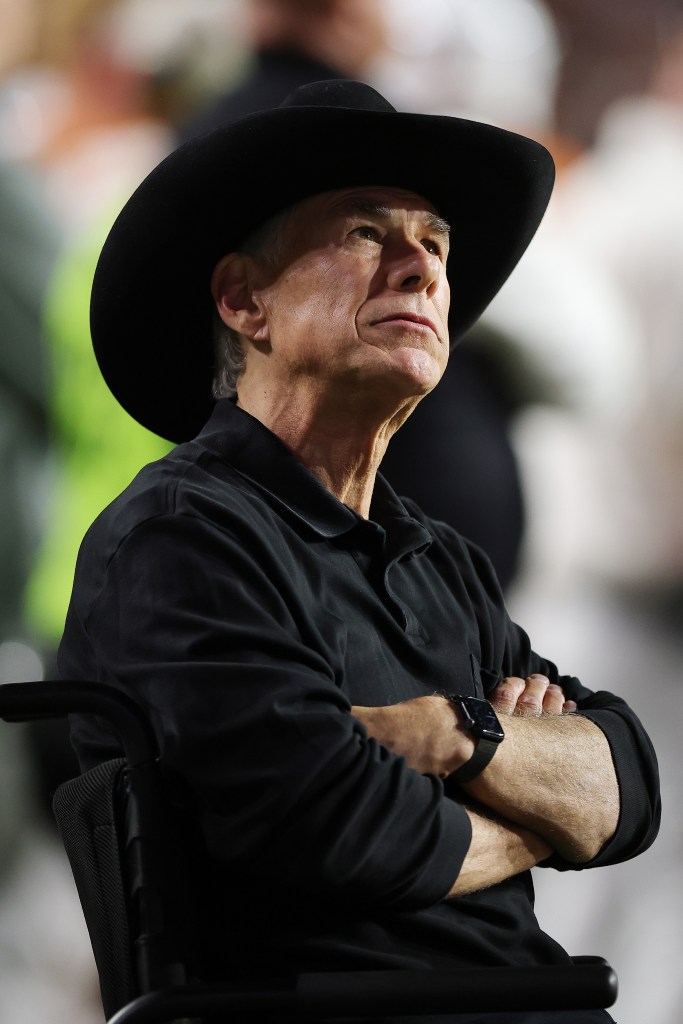 Texas Governor Greg Abbott in a black cowboy hat and shirt, seated in a wheelchair with arms crossed.