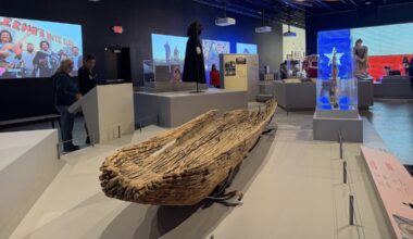 A view of a gallery exhibit with a hand-hewn aged wooden canoe in foreground.