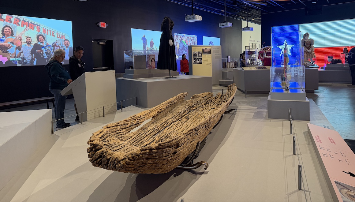 A view of a gallery exhibit with a hand-hewn aged wooden canoe in foreground.