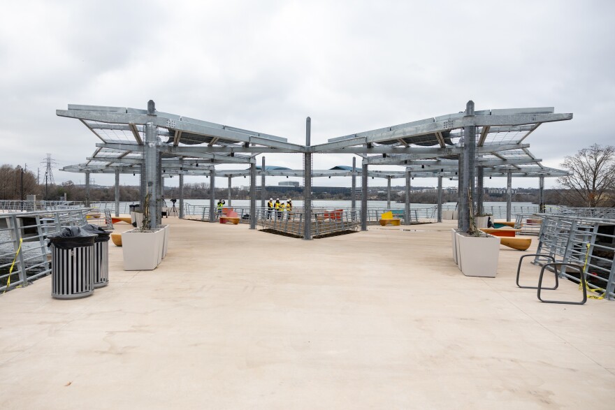 A view of the bridge from the plaza in the middle. Large steel shade structures will support vines overhead. Planters, bike rans and trash cans are visible. Some people in hard hats and high-visibility vests are in the background.
