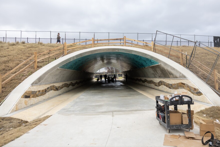 A view of the pedestrian underpass beneath Pleasant Valley Road. A man is seen walking on the sidewalk above. Inside the tunnel is a cluster of people working to install the mosaic tile mural.