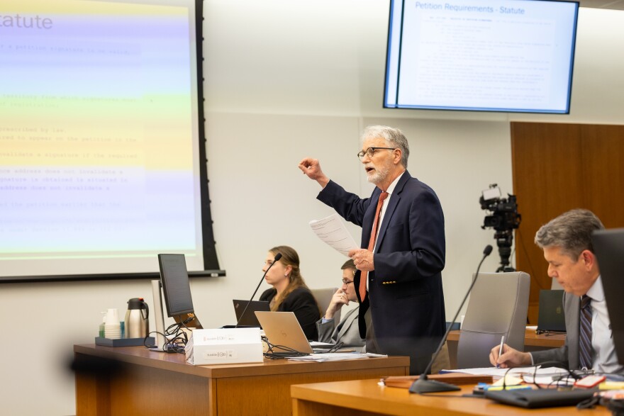 A man stands at a table with screens behind him. There are others at the table and he is gesturing. 