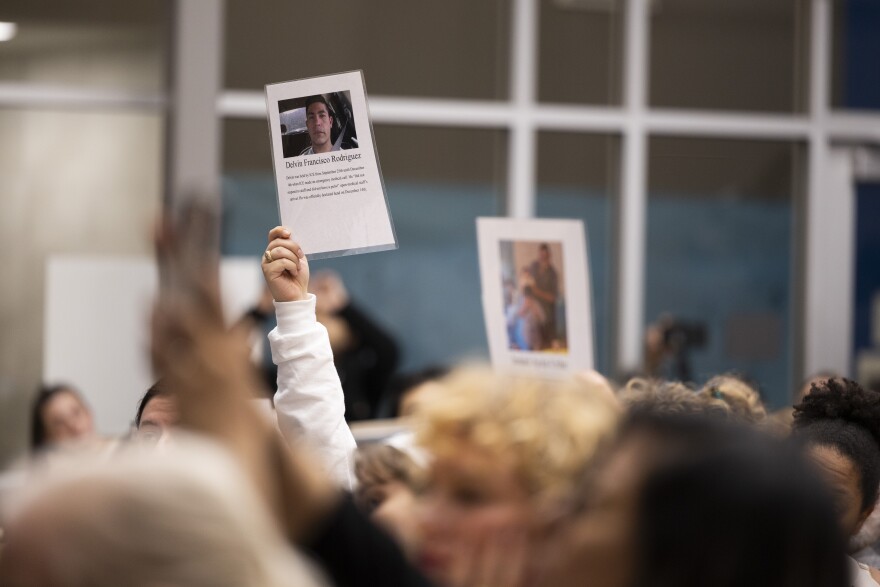 A photo is held up above a crowd showing the image of man who text under the photo identifies as someone taken by ICE.