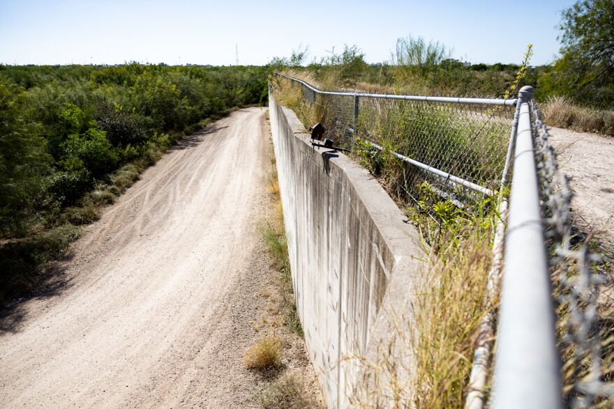 A dirt road is shown at left with a concrete wall and fence at right.