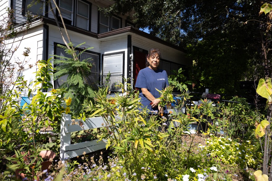 A woman stands in the front yard surrounded by plants.