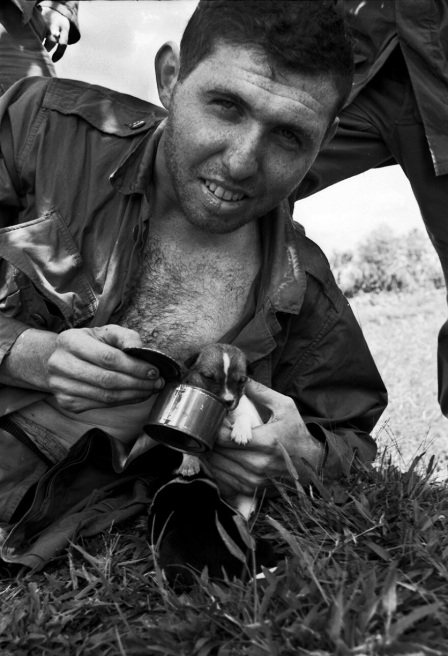 A black and white photo of a soldier with his uniform shirt open, feeding a tiny puppy out of a can of army rations.