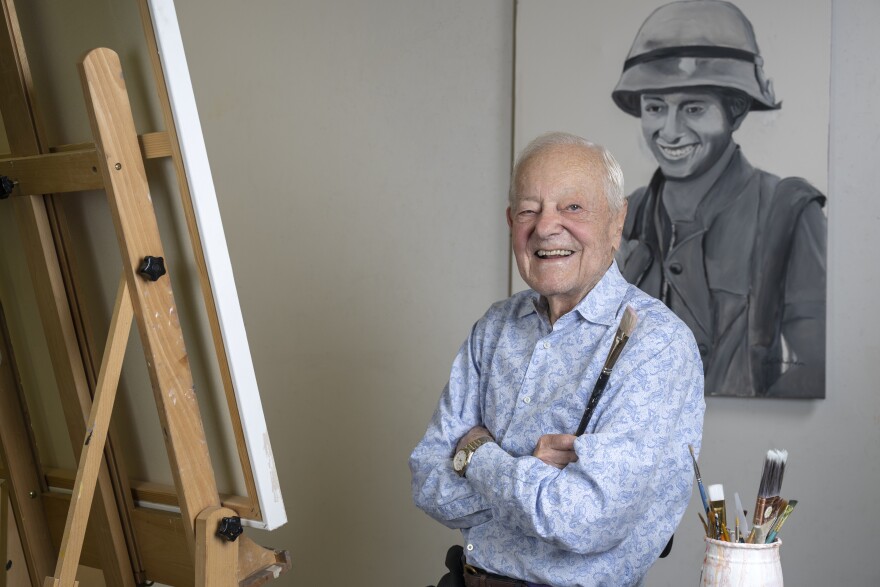 A photo of Bob Schieffer, an elderly white man in a blue patterned shirt, sitting in front of an easel and holding a paintbrush. A painting of a smiling soldier hangs on the wall behind him.