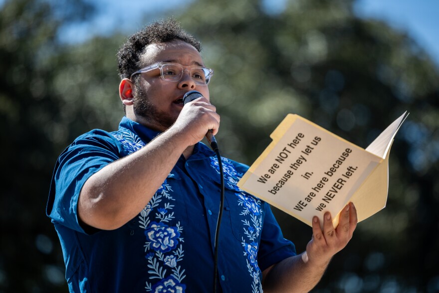 A young man holds up a folder as he speaks into a microphone. The folder reads "We are not here because they let us in. We are here because we never left." 