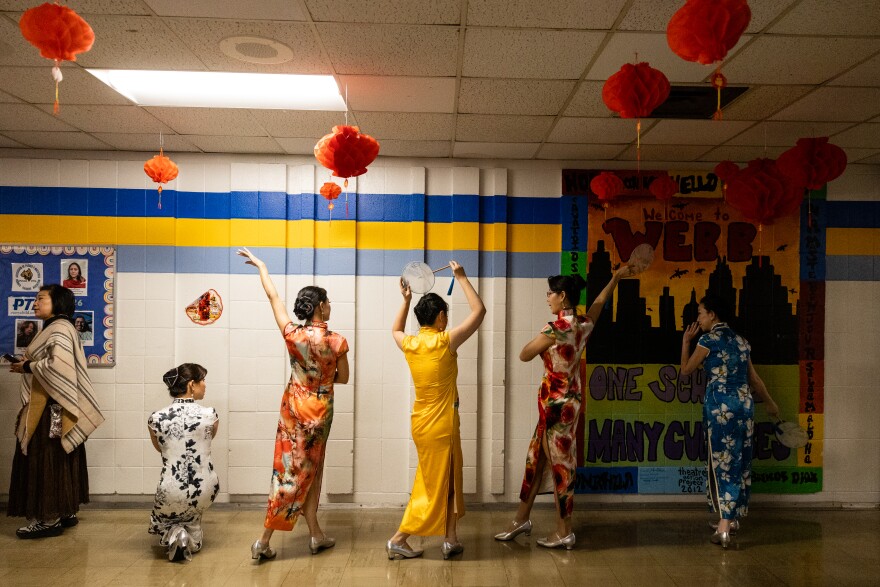 Women in traditional Chinese dress stand near a wall, with their backs to the camera, rehearsing a dance.