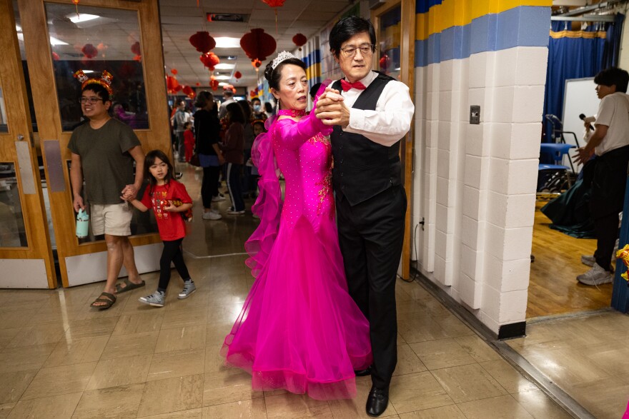 An older woman in a small crowd and bright pink dress dances with a man in a formal suit in a hallway at a school.