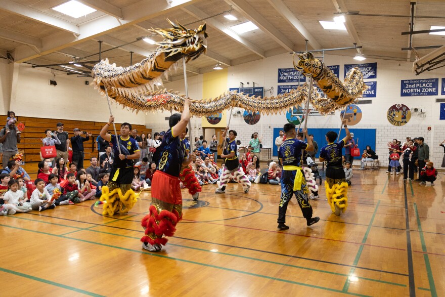 Men hold up sticks with a dragon attached at the top as they dance in front of young children.