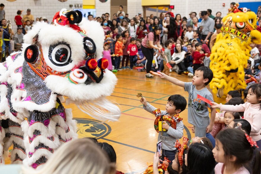 A crowd of children raise envelopes as performers wearing a dragon costume approaches them.