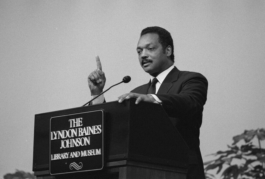 A man stands at a podium and speaks while pointing his hand. The photo is in black and white. 