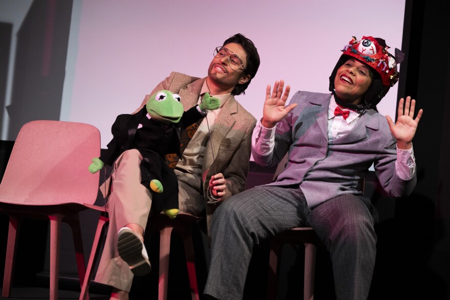 Two performers sit on stage, one of the holding a kermit the frog puppet. The other is wearing a red hat with an eye ball on the front.