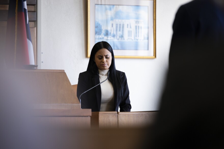 A woman on a courthouse witness stand looks down. 