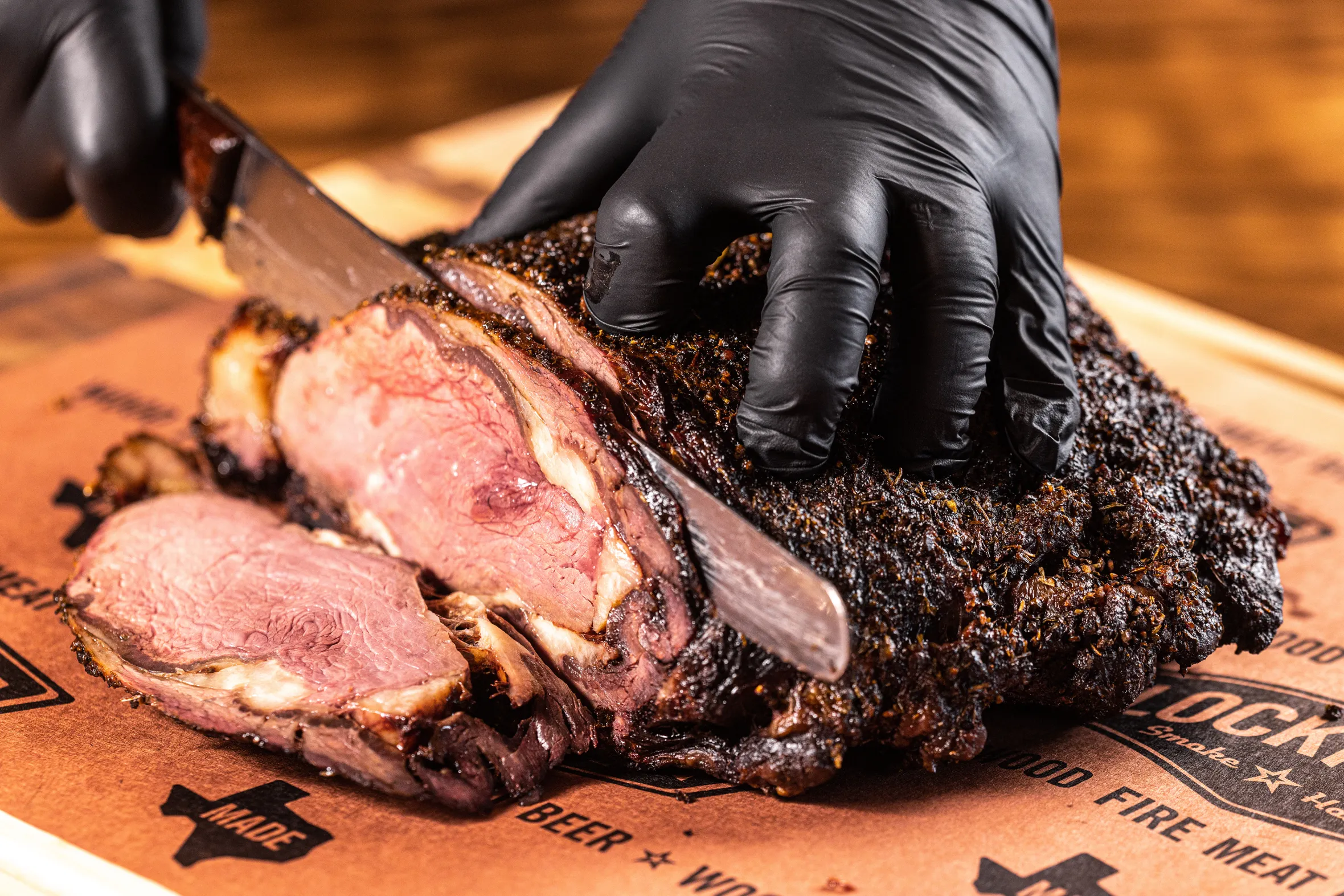 A close-up of a black-gloved hand slicing a large piece of Lockhart BBQ, revealing the pink meat inside.