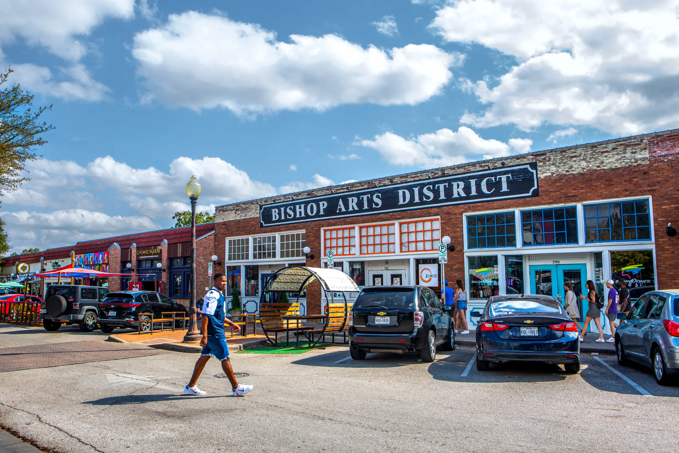 Street view of the Bishop Arts District, Dallas, with a large "BISHOP ARTS DISTRICT" sign on a brick building, and people walking along the sidewalk.