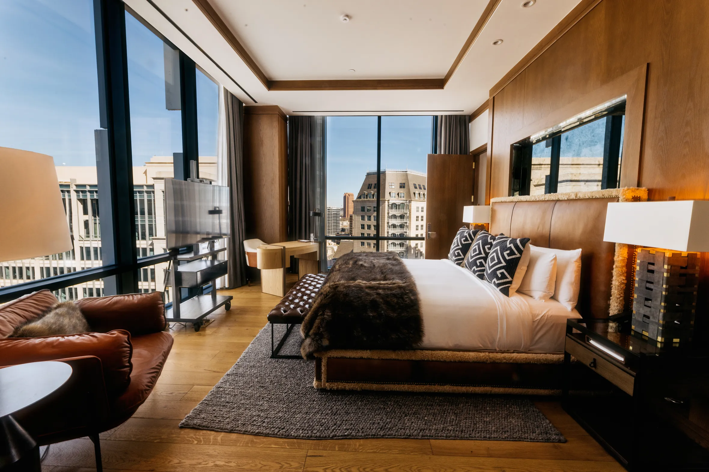 A hotel room at Hotel Swexan with floor-to-ceiling windows, a large bed with a fur throw, a brown leather armchair, and a modern desk.